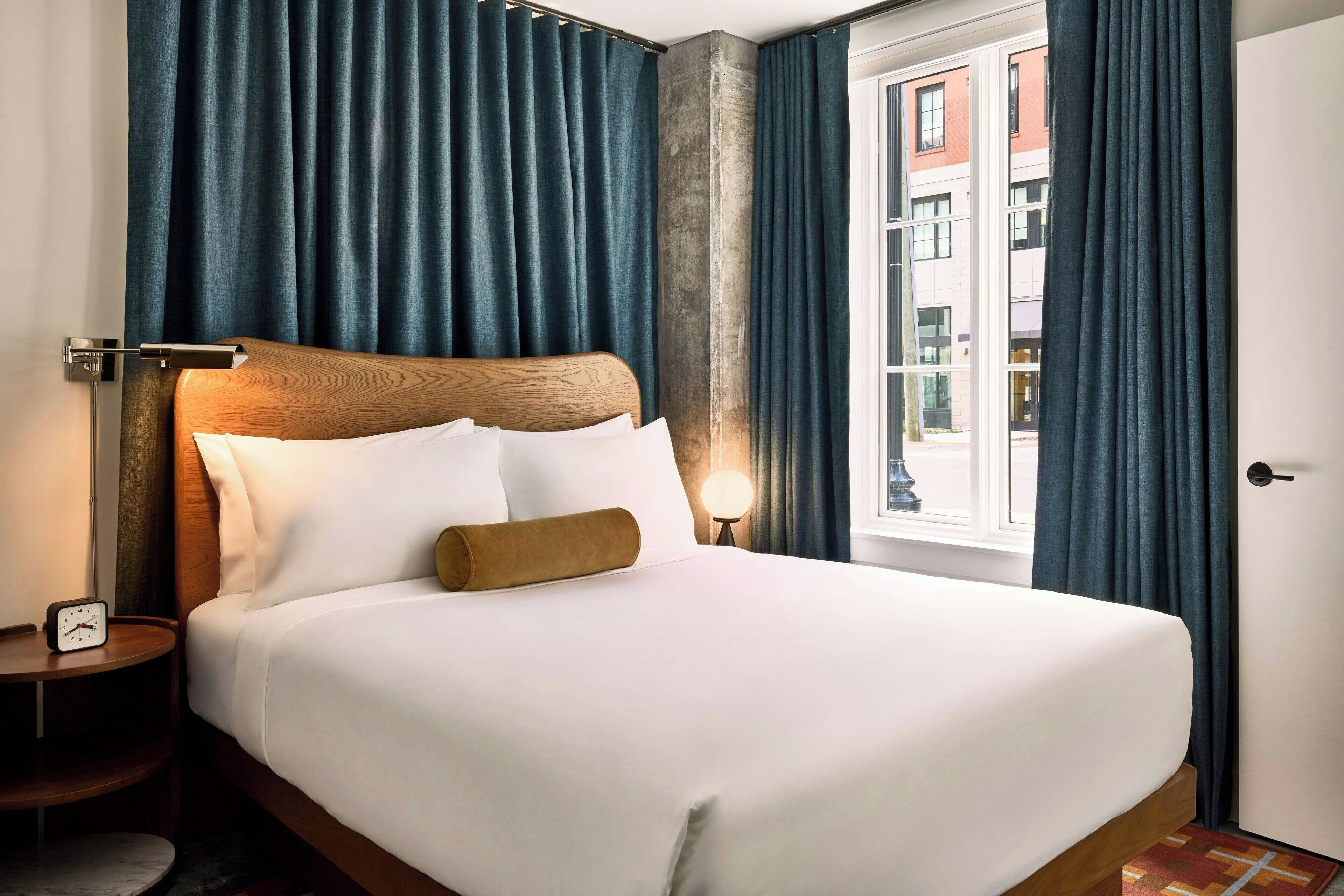 Una cama bien hecha con sábanas blancas y un cabecero de madera, una mesita de noche con una lámpara, y vistas de un edificio a través de una ventana con cortinas azules.