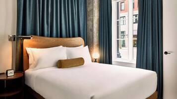 Una cama bien hecha con sábanas blancas y un cabecero de madera, una mesita de noche con una lámpara, y vistas de un edificio a través de una ventana con cortinas azules.