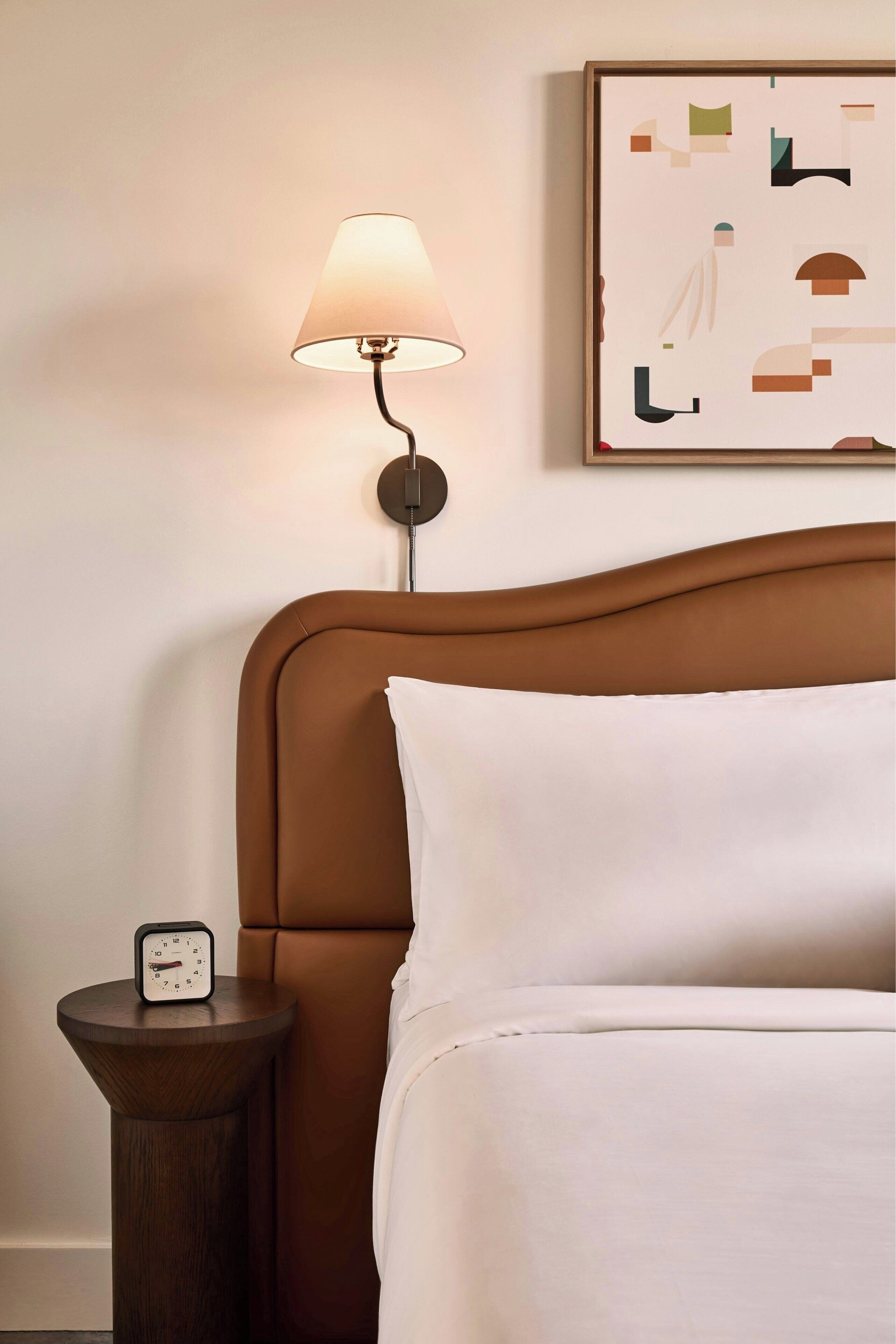 Ein ordentlich bezogenes Bett mit weißen Leinen, ein braunes Kopfteil, ein Nachttisch mit einer Uhr, eine Wandlampe und ein gerahmtes abstraktes Kunstwerk.