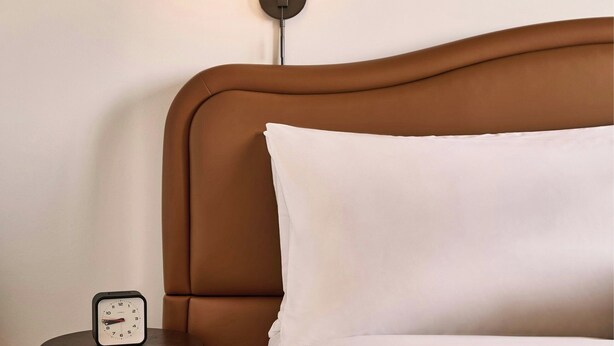 A neatly made bed with white linens, a brown headboard, a bedside table with a clock, a wall-mounted lamp, and a framed abstract artwork. - The Ann Savannah, Apartments By Marriott Bonvoy (Savannah)