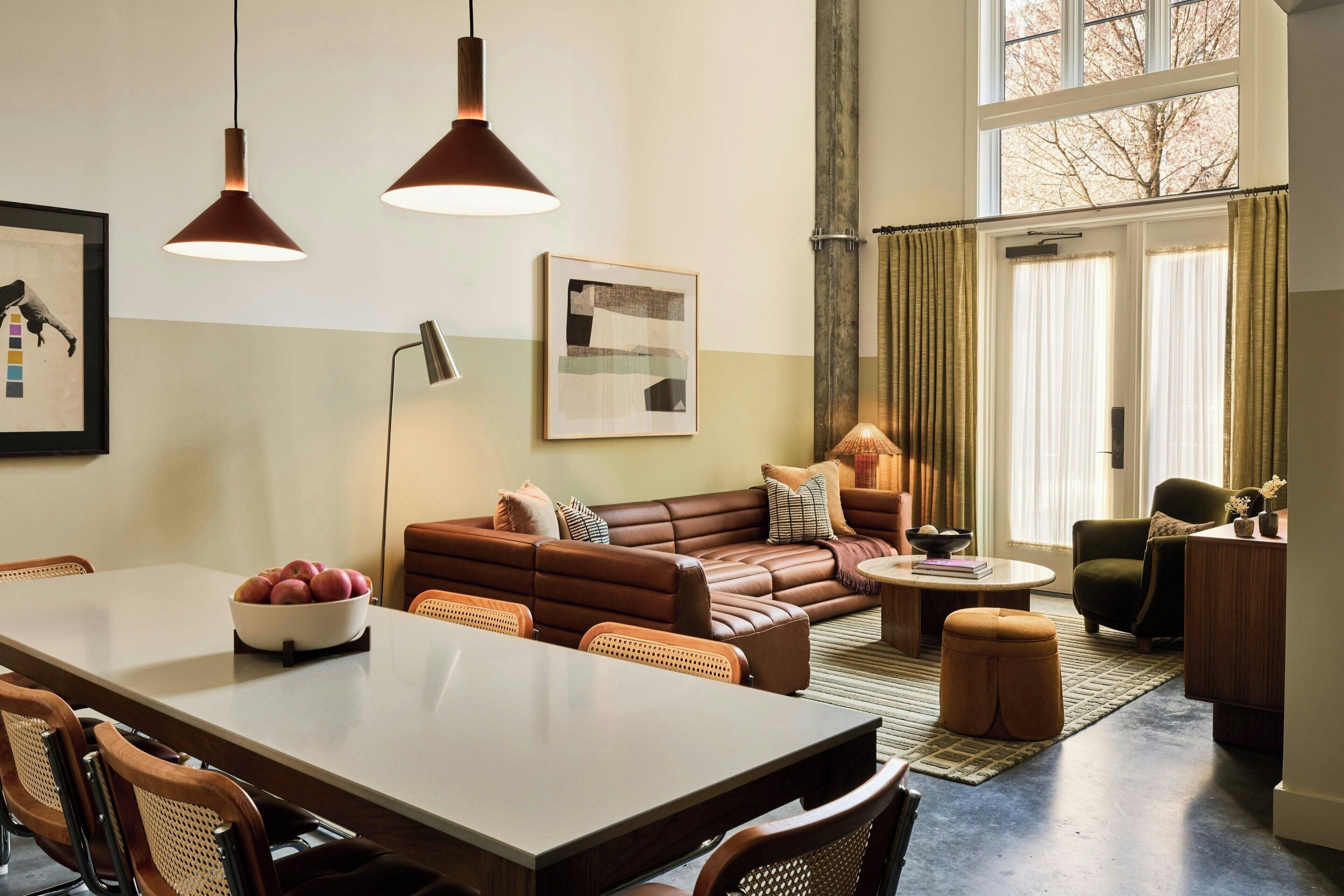A spacious living room with a brown leather sofa, a dining table with chairs, and a large window with curtains.