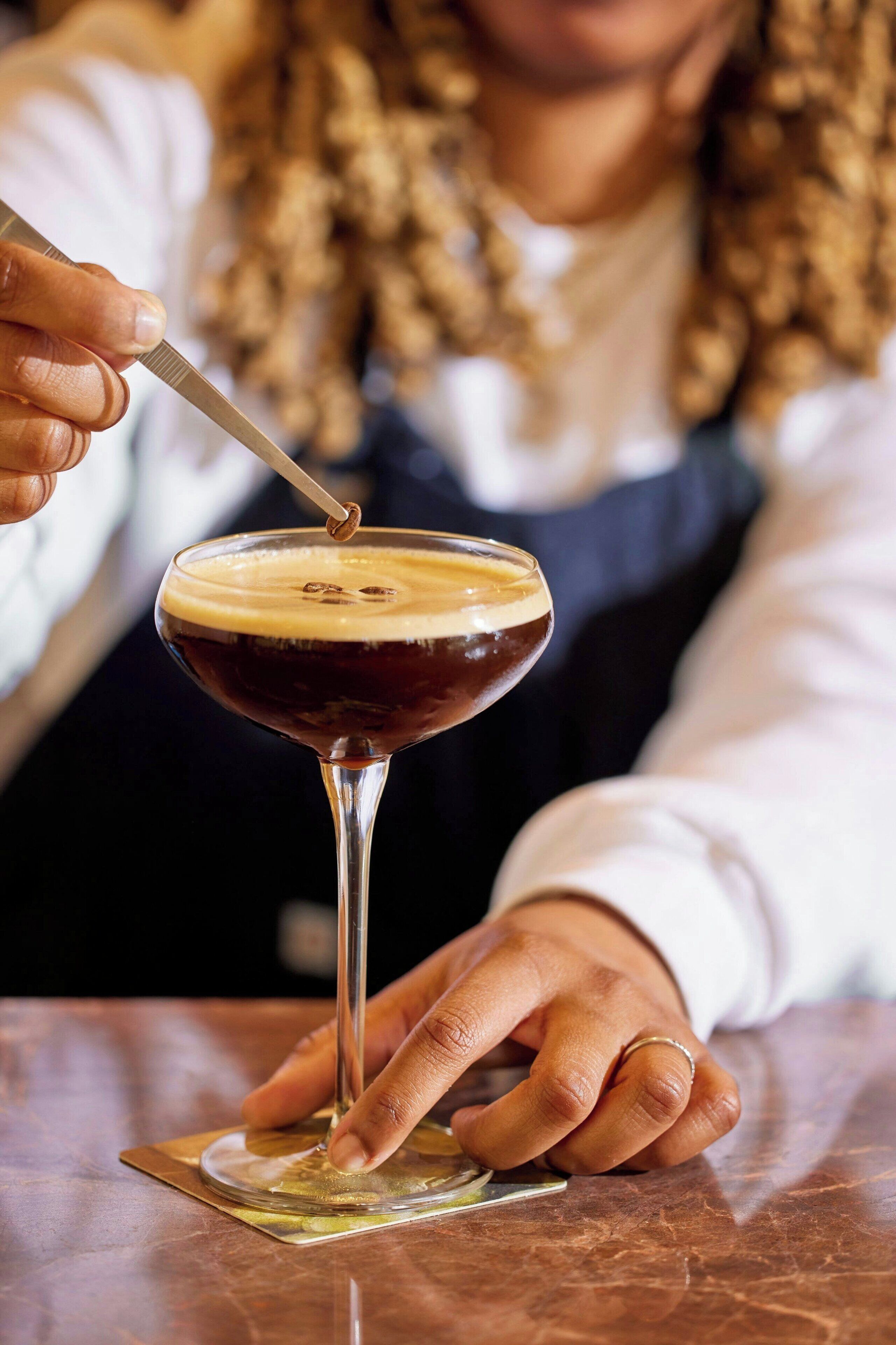 A person holding a cocktail glass with a spoon, a bar mat, and a bar counter.