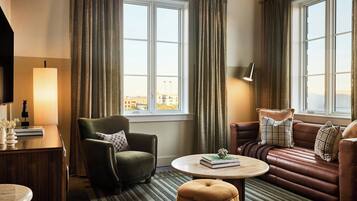 A hotel room with a green armchair, a brown leather sofa, a round coffee table, and a view of the city through large windows.