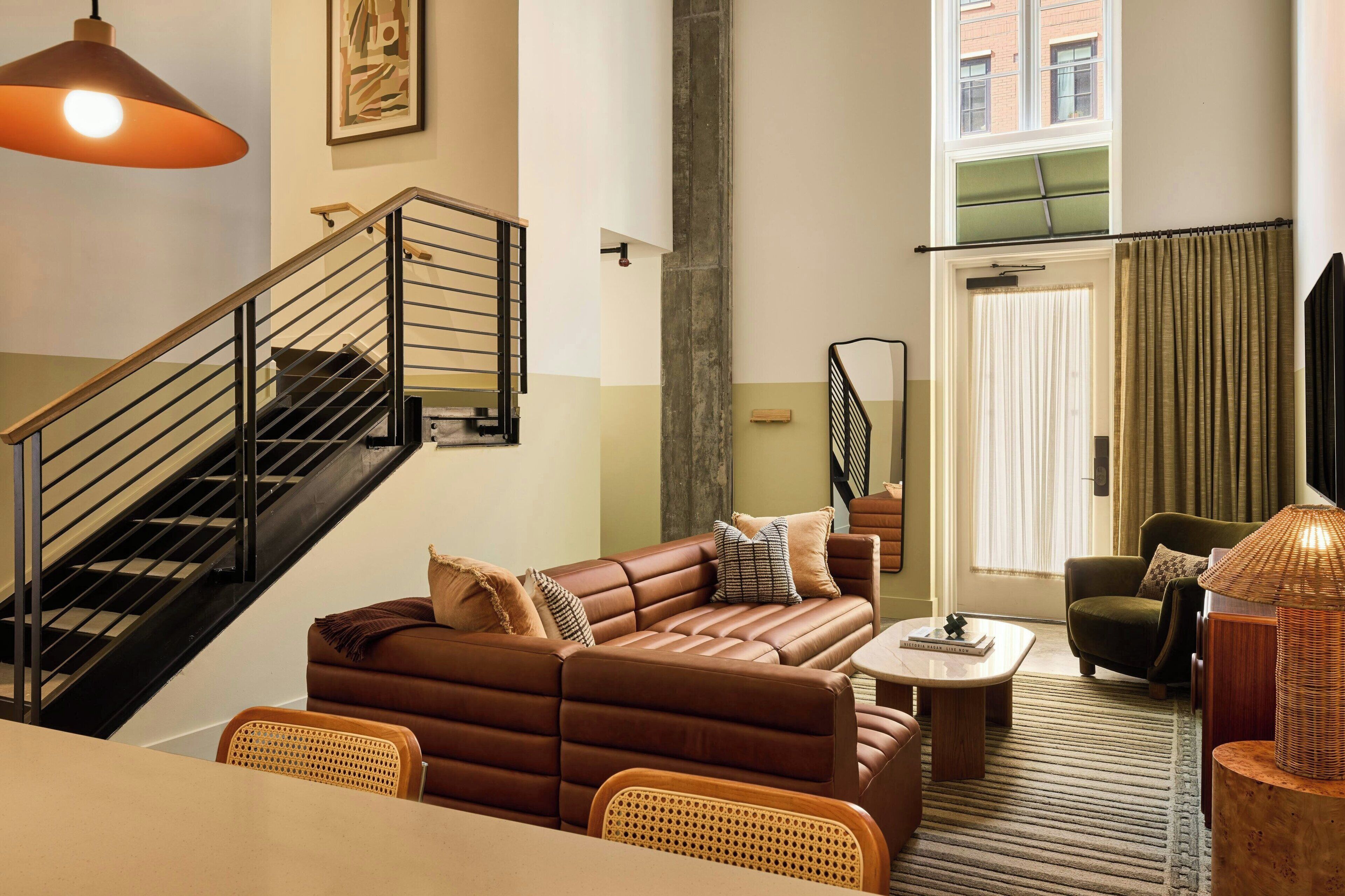 Un salon moderne avec un canapé en cuir marron, une table à manger, des chaises en osier, un escalier avec des rampes noires et une grande fenêtre avec des rideaux.