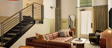 A modern living room with a brown leather sofa, a dining table, wicker chairs, a staircase with black railings, and a large window with curtains.