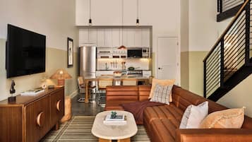 A modern living room with a sofa, coffee table, and a TV. There is a kitchen in the background with cabinets and appliances.