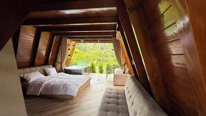 A cozy attic bedroom with a wooden sloped ceiling, a large bed, a bench, and a view of greenery outside. - Patate Lodge (Patate)