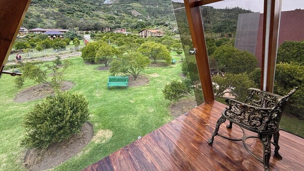 A room with a large window overlooking a landscaped garden with a bench and trees. - Patate Lodge (Patate)