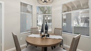 A dining area with a round table, four chairs, and two large windows with blinds.