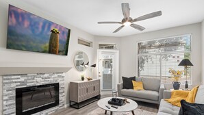 A living room with a fireplace, a ceiling fan, and a large window with blinds.