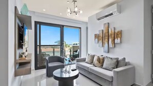 A modern living room with a grey sofa, a coffee table, and a TV mounted on the wall. There are large windows providing a view of the outside.