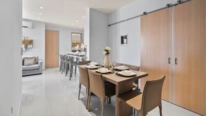 A modern dining area with a long table set for six, surrounded by chairs. There is a sofa and a dining table in the background.
