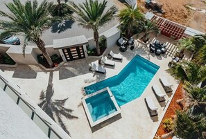 An aerial view of a resort with a swimming pool, lounge chairs, palm trees, and a beach.
