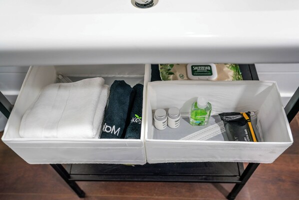 Baño con lavamanos blanco y dos cajones de almacenamiento. Un cajón contiene una toalla blanca, una toalla oscura y una botella de champú. El otro cajón tiene una pastilla de jabón, una botella de loción y otra pastilla de jabón.