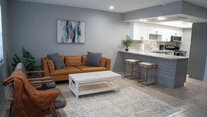 Ein modernes Wohnzimmer mit einer Couch, einem Couchtisch und Barhocker. Es gibt eine Küchenzeile mit Spüle, Herd und Mikrowelle.