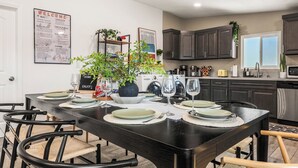 Un comedor con una mesa de madera oscura dispuesta para seis personas, rodeada de sillas negras. Un letrero de bienvenida está en la pared, y hay una planta en maceta sobre la mesa.