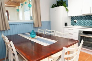 A dining area with a wooden table, white chairs, a blue vase, and a kitchen with a white refrigerator and stove. - Beachfront apartment with pool, 4 minutes walk to Indian Ocean. Sleeps 6 (Quinns Rocks)