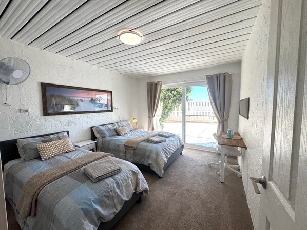 A room with two beds, a ceiling fan, a table with a lamp, and a view of the outdoors through a sliding door. - Beachfront apartment with pool, 4 minutes walk to Indian Ocean. Sleeps 6 (Quinns Rocks)