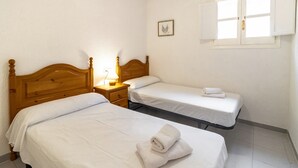 A room with two single beds, a nightstand with a lamp, a window with shutters, and a small framed picture on the wall.