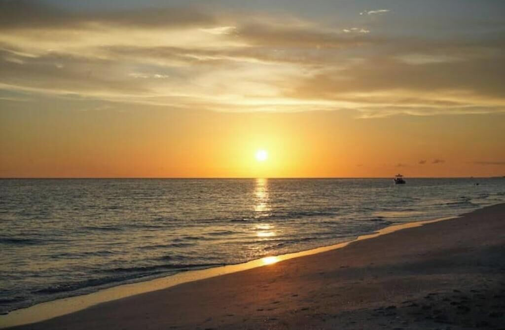 Solnedgang over en strand med roligt vand og en båd i det fjerne.