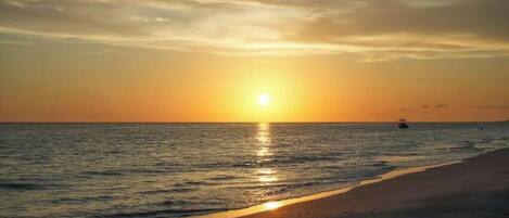 Solnedgang over en strand med roligt vand og en båd i det fjerne.