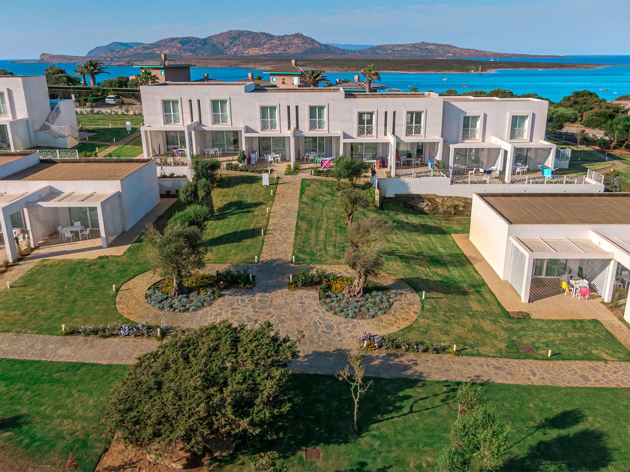 Un complexe hôtelier moderne composé de plusieurs bâtiments blancs, avec une cour centrale dotée d’un sentier en pierre, et offrant une vue sur la mer et les montagnes en arrière-plan.
