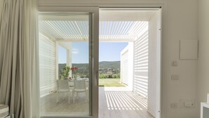 A room with a sliding glass door leading to an outdoor patio with a table and chairs, a view of a green landscape, and a white wall with a light switch. - Capo Falcone Charming Apartment by Interhome (Stintino)