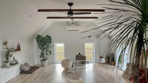 A spacious room with a high ceiling, wooden beams, a ceiling fan, and a large window providing natural light.