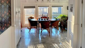 A dining area with a round table, chairs, and a chandelier. There are large windows and a potted plant.