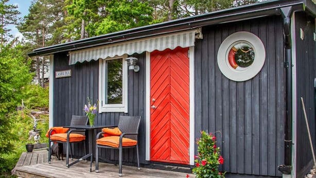 Une cabane avec une porte rouge, un petit porche avec deux chaises et une table, et une plante en pot.