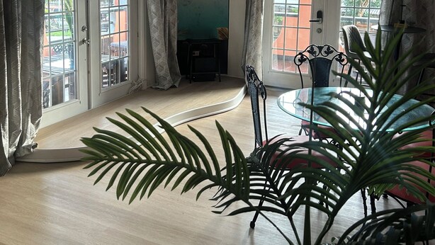 A hotel room with a ceiling fan, a TV mounted on the wall, a large window with curtains, a dining table with chairs, and a potted plant.