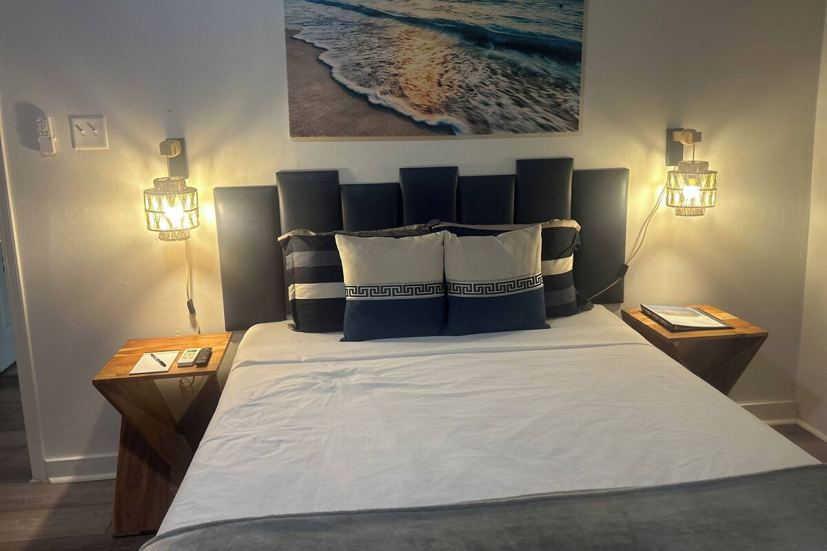 A bedroom with a large bed, a headboard with multiple pillows, two bedside tables with lamps, and a wall-mounted painting of a beach scene.