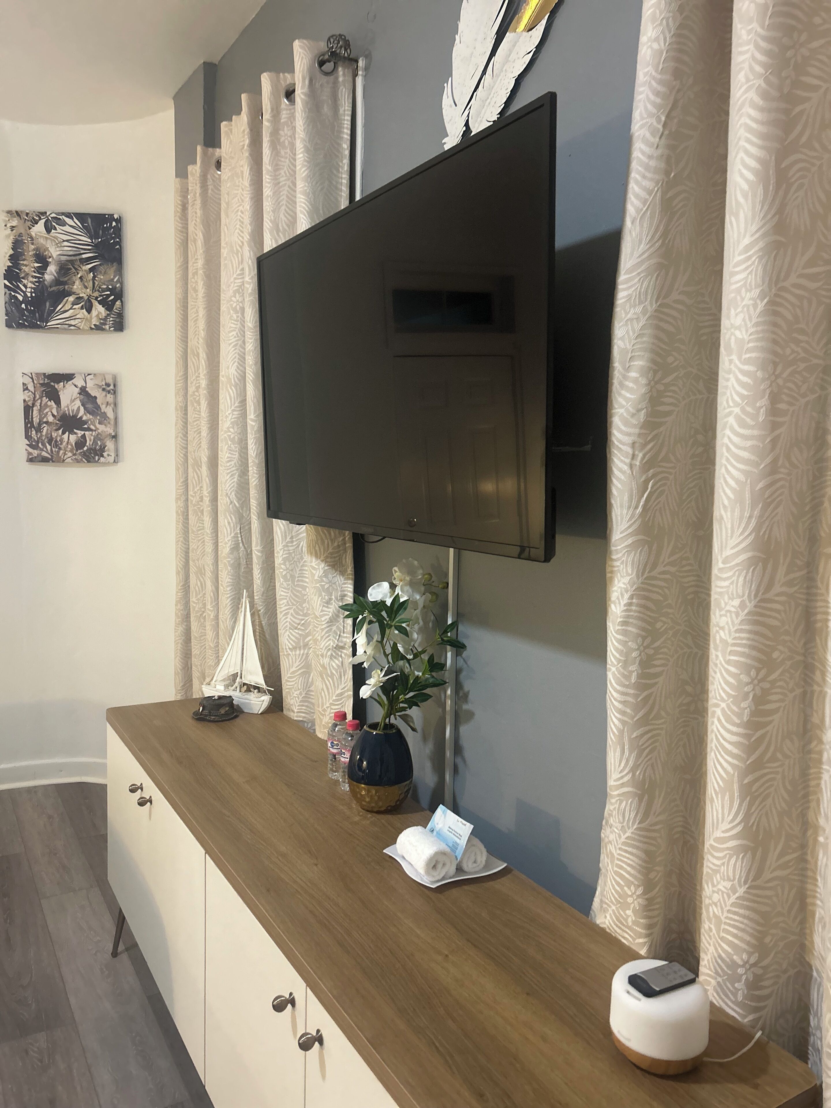 Ein modernes Wohnzimmer mit einem flachbildfernseher an der Wand, einem Holztischchen mit Dekoration und gemusterten Vorhängen.
