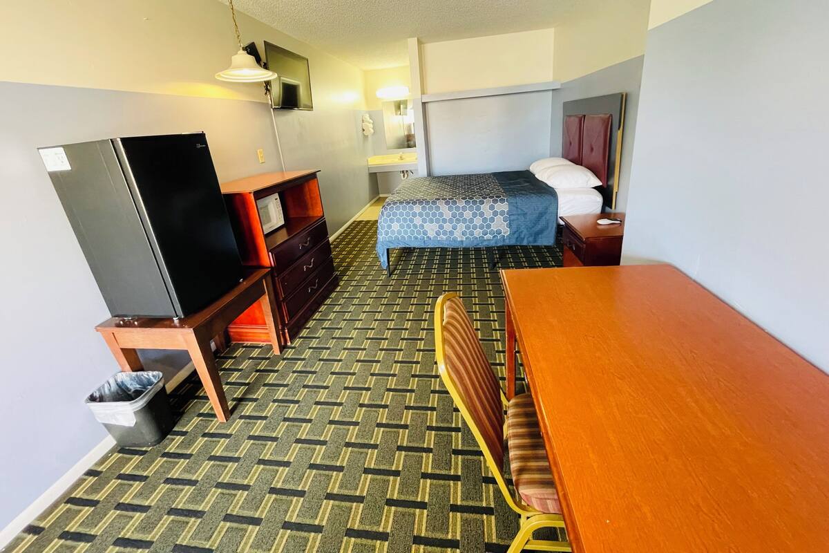 A compact hotel room with a bed, a desk, a chair, a dresser, a microwave, and a television.