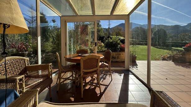 Ein Wintergarten mit Esstisch und Rattanmöbeln, mit Blick auf einen Garten und Berge.