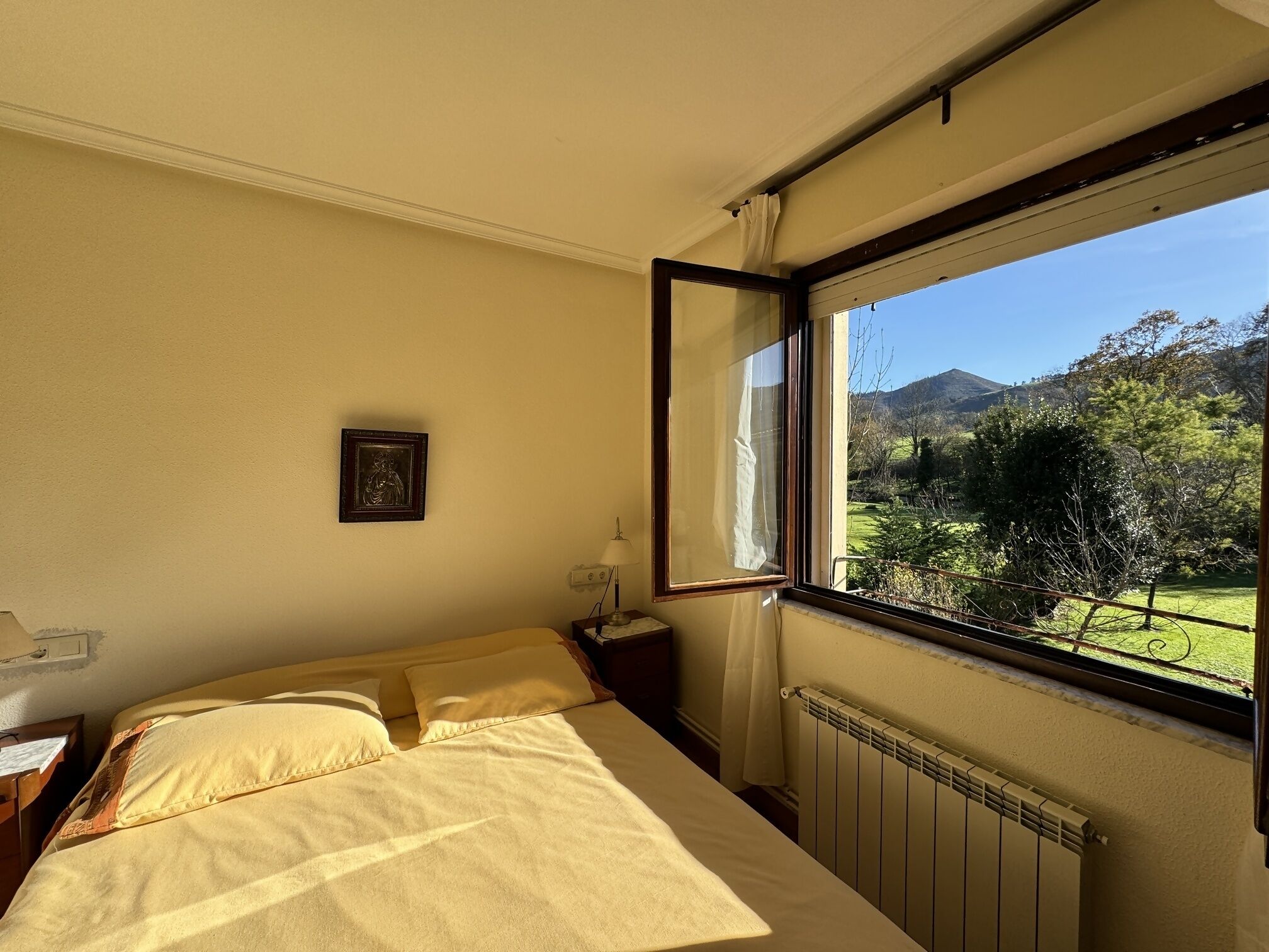 Un dormitorio con cama, mesita de noche, lámpara, cuadro enmarcado y ventana con vistas a un paisaje verde y montañas.
