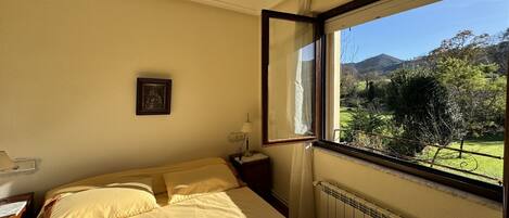 Un dormitorio con cama, mesita de noche, lámpara, cuadro enmarcado y ventana con vistas a un paisaje verde y montañas.