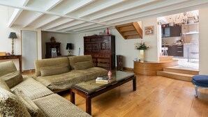 A spacious living room with a high ceiling, wooden flooring, a sofa set, a coffee table, and a view into the kitchen area.