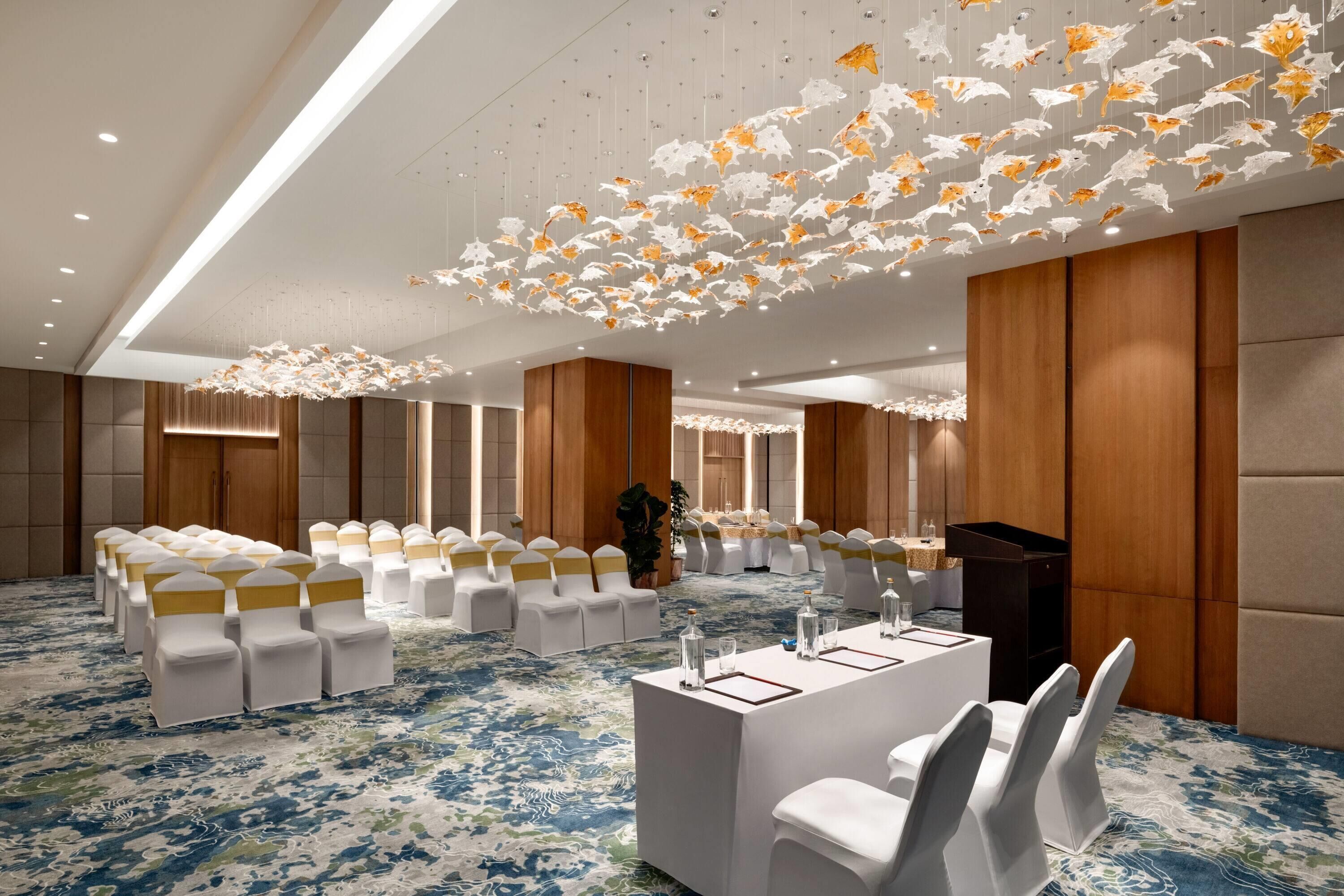 Une salle de conférence avec des rangées de chaises blanches, une longue table avec des verres d’eau et un plafond orné de décorations suspendues.