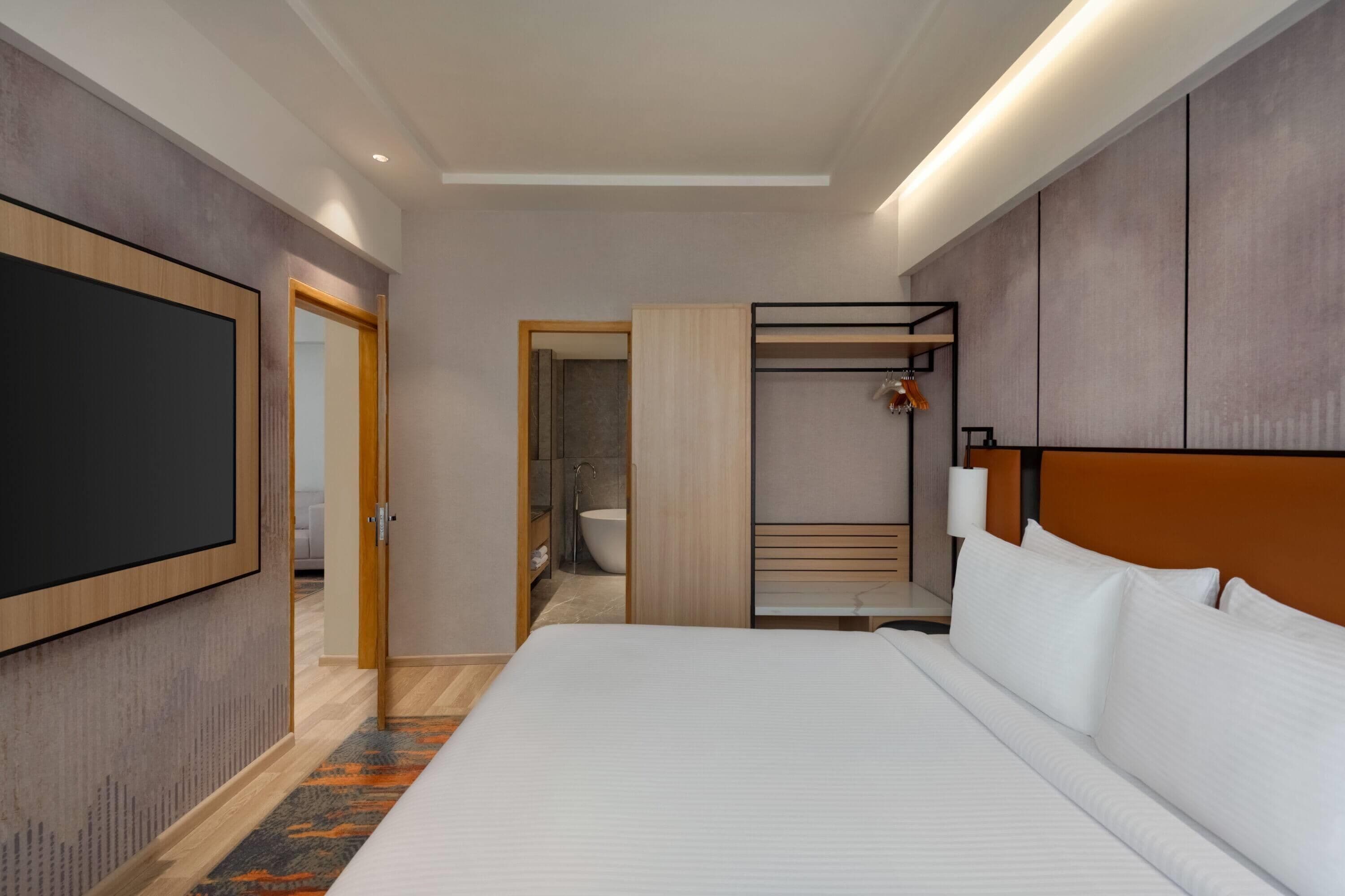 Una habitación de hotel moderna con una cama grande, televisión, vestidor y un baño visible a través de una puerta abierta.