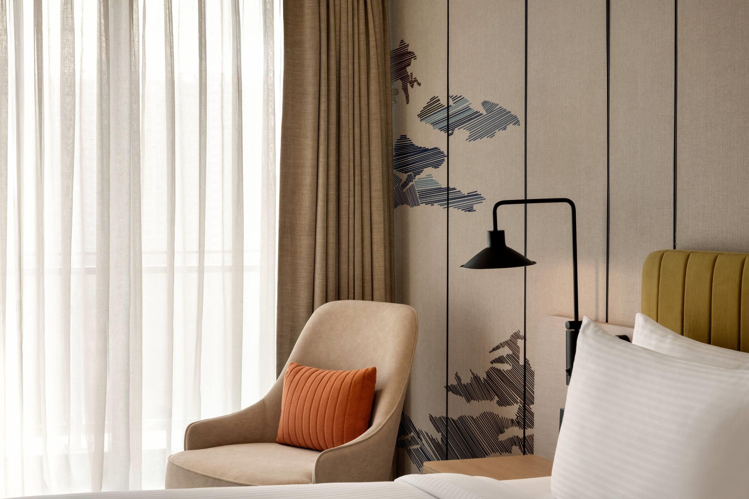 Een hotelkamer met een bed, een stoel, een lamp en een raam met gordijnen.