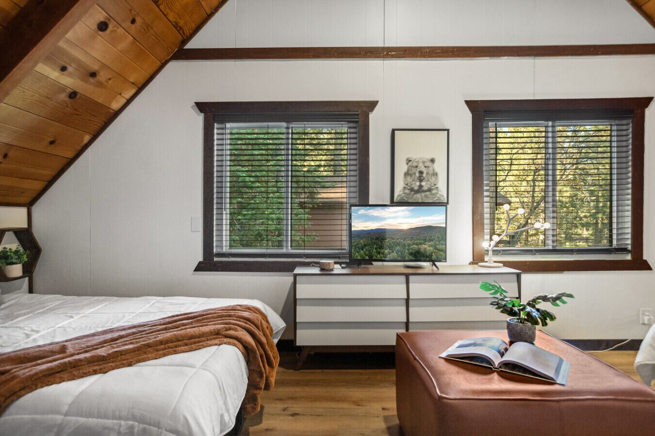 Une chambre avec un plafond en pente, un lit recouvert d’une couverture marron, un bureau avec un ordinateur et une plante, et deux fenêtres équipées de stores.