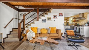 A cozy living room with a wooden staircase, a leather sofa with yellow cushions, a coffee table, and a kitchen area visible through an open door. - TreeLicious | Jacuzzi, Cozy Chalet close to Slopes (Big Bear Lake)