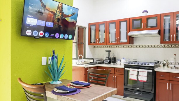 A kitchen with a TV mounted on the wall displaying a movie, wooden cabinets, a stove, and a dining table with chairs. - 27 Ely House (Cancún)