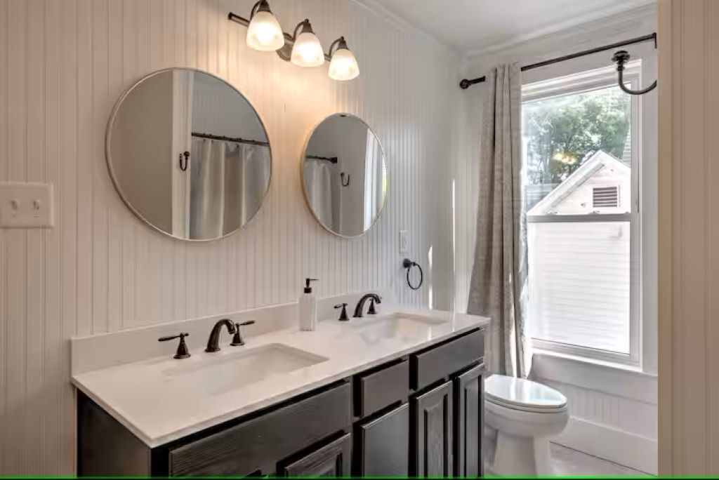 Un baño con dos lavamanos, inodoro y ventana con cortinas.