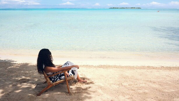 A person sitting on a wooden chair on a sandy beach with clear turquoise water and a distant island. - Single Tropical Pool Villa (MOOREA MAIAO)