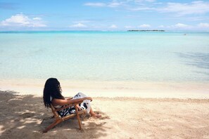 Una persona sentada en una silla de madera en una playa arenosa con agua turquesa cristalina y una isla distante.