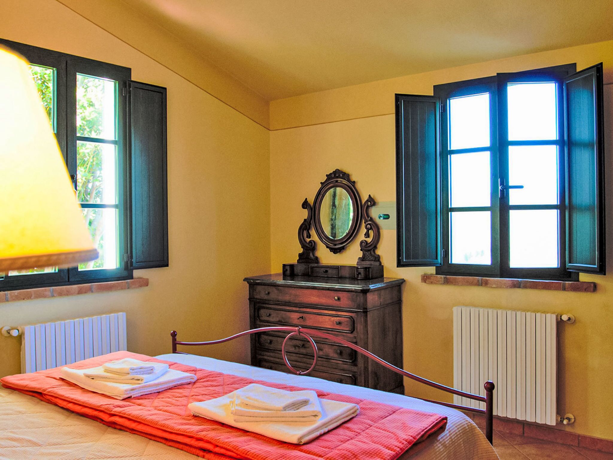 Une chambre à coucher comprenant un lit, une commode, un miroir et deux fenêtres équipées de volets.