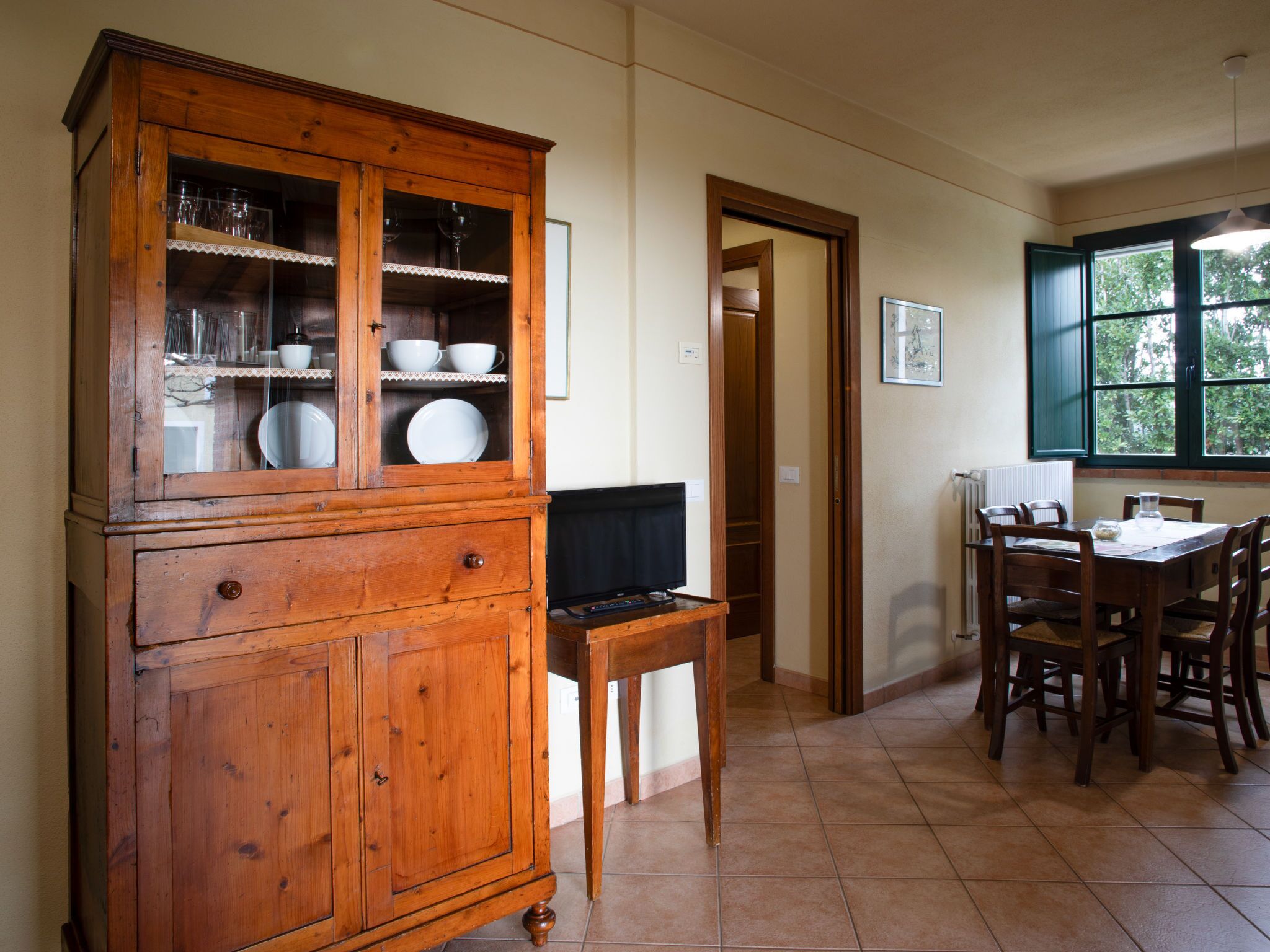 A wooden cabinet with glass doors, a dining table with chairs, and a window with a view of trees.