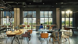 A spacious indoor area with multiple tables and chairs, large windows offering views of the outside, and a person working at a desk. - Moxy Warsaw City (Warsaw)